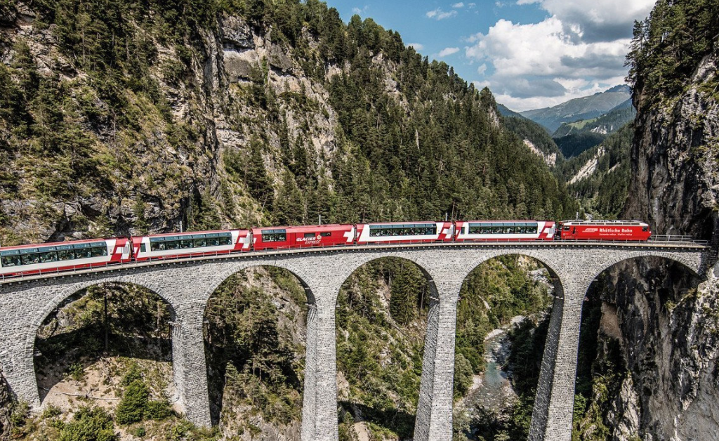 Bernina Express train crossing the iconic Landwasser Viaduct, surrounded by lush green forests and rugged Swiss Alps under a sunny sky.