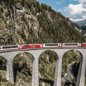 Bernina Express train crossing the iconic Landwasser Viaduct, surrounded by lush green forests and rugged Swiss Alps under a sunny sky.
