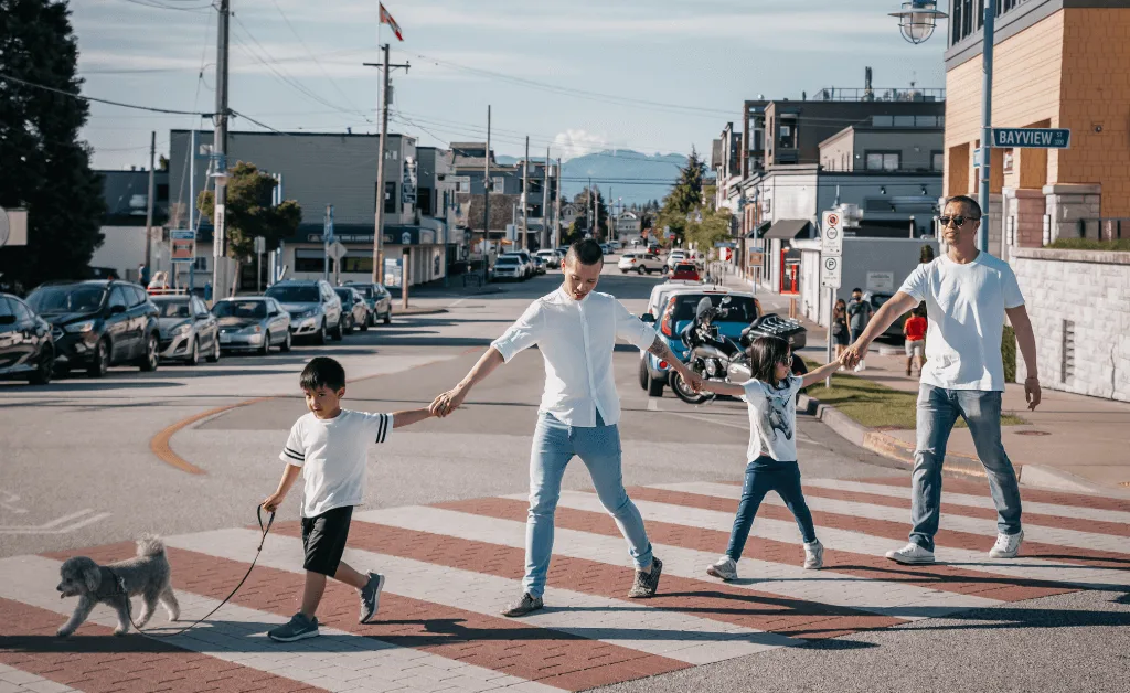 A family of four and their small dog crossing a pedestrian crosswalk in a bustling urban area, with cars and buildings in the background.