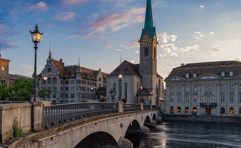 A scenic sunset view of Zurich, Switzerland, featuring the Fraumünster Church, a historic bridge, and the Limmat River.