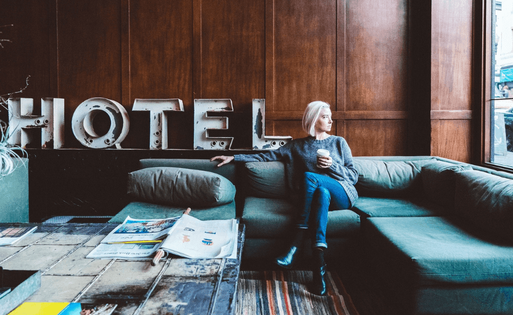 Woman relaxing on a couch in a hotel lobby, drinking coffee and looking out the window. Hotel sign behind her.