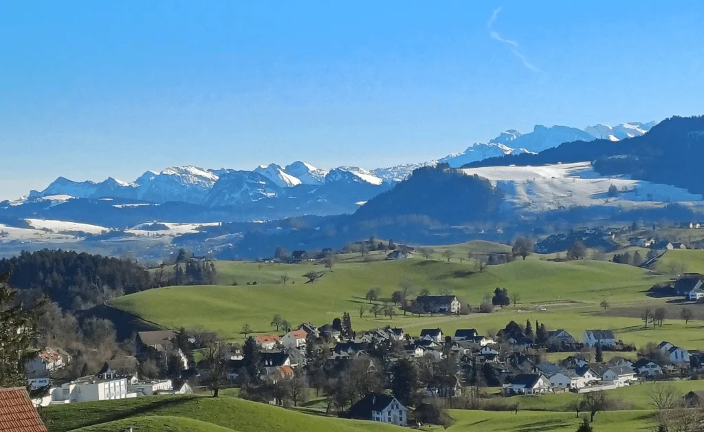 Panoramic view from a balcony overlooking the rolling green hills and snow-capped peaks of the Glarner Alps in Switzerland.