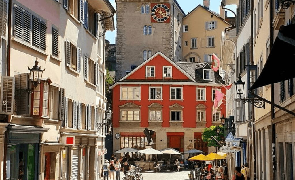 A view down a cobblestone street in Zürich's Old Town (Altstadt) on a sunny day, flanked by tall, multi-story historic buildings with shutters. In the center is a bright red, three-story building with white-trimmed windows, behind which a tall, stone clock tower with a large red and gold clock face rises. People are seated at an outdoor café with umbrellas in the foreground.