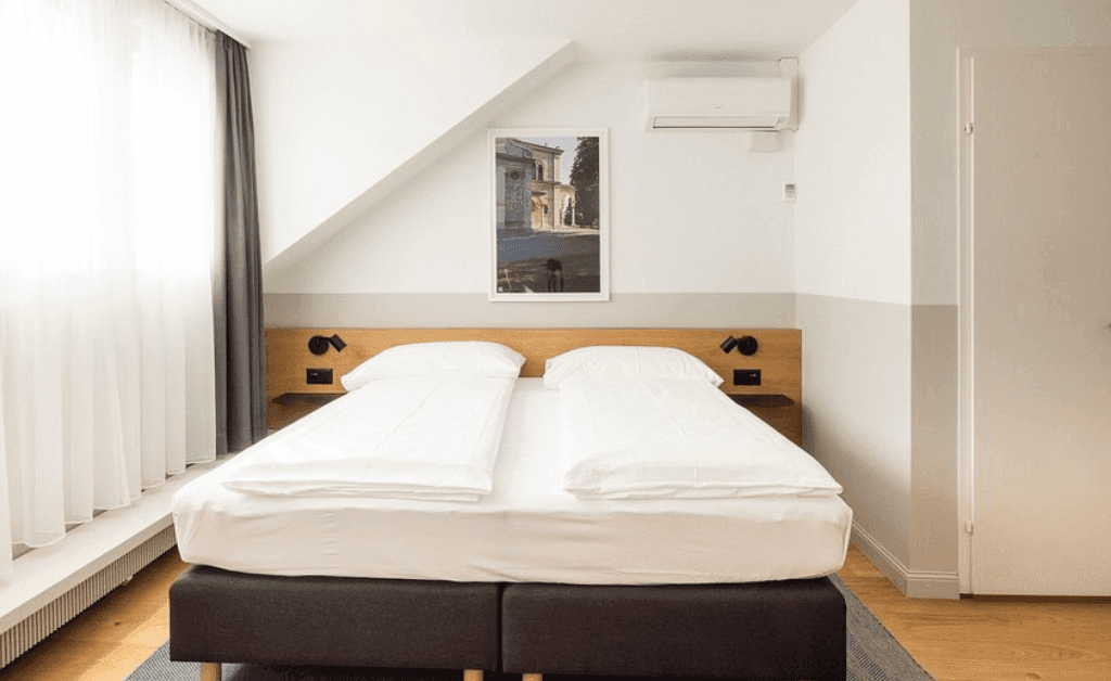 Minimalist room interior at Fred Hotels Zürich with a double bed, air conditioning, and a slanted ceiling with white and grey walls.