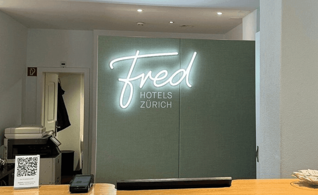 Reception desk at Fred Hotels Zürich featuring a modern white neon script sign on a light green wall.