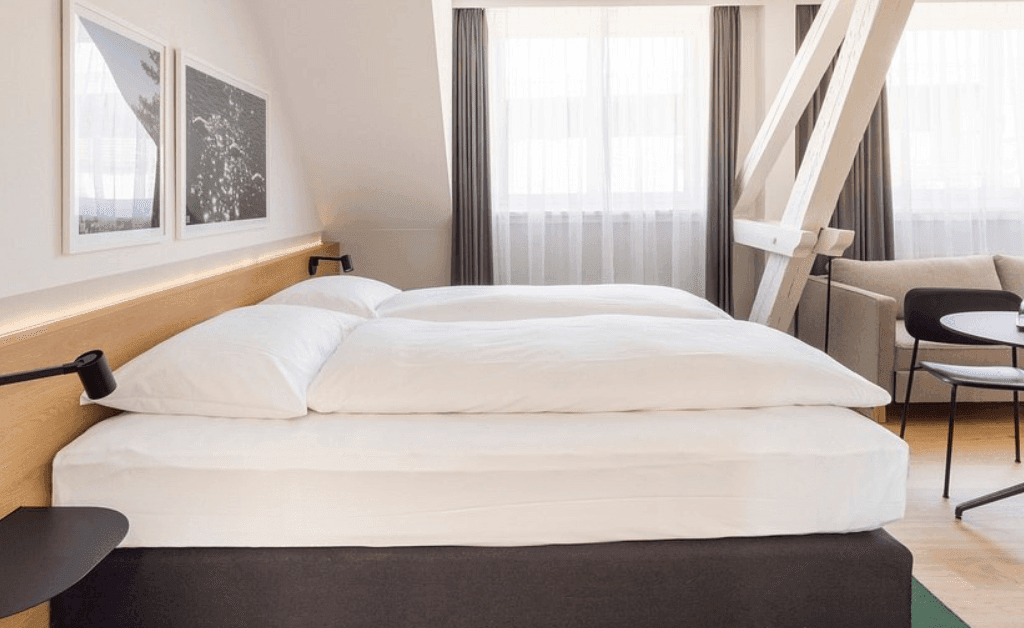 Modern double room at Fred Hotels Zürich featuring a neutral color palette, wooden headboard, exposed structural beams, and a small sitting area.