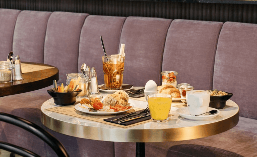 A table with the Ruby Mimi Hotel’s healthy breakfast buffet, including eggs, juice, coffee, granola, and pastries next to a mauve velvet sofa.