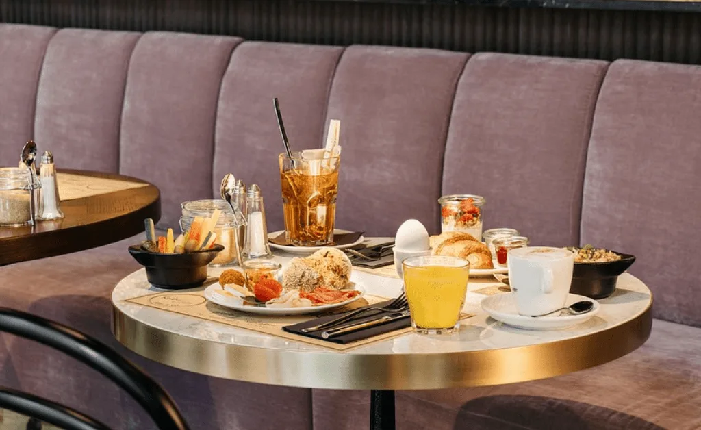 A table with the Ruby Mimi Hotel’s healthy breakfast buffet, including eggs, juice, coffee, granola, and pastries next to a mauve velvet sofa.