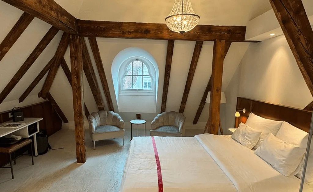 Interior of a Ruby Mimi Hotel Loft Room in Zürich featuring exposed wooden ceiling beams, an arched window, and a white queen bed.