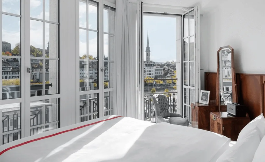 Ruby Mimi Hotel room with balcony view of Zürich's Limmat river, historic buildings, and the Grossmünster church spire.