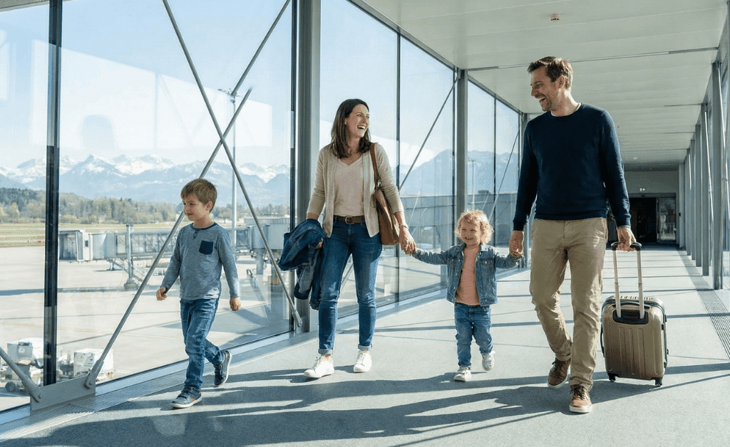 A family walking through a modern glass bridge at Zurich Airport toward the check-in area.