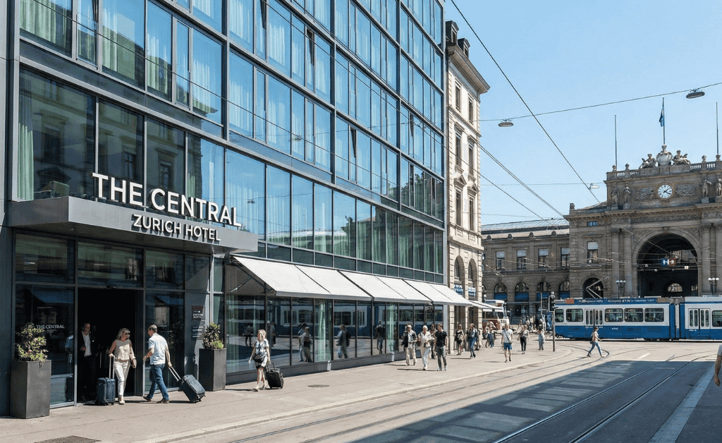 Sunny daytime view of a modern hotel with a glass facade located directly opposite the historic Zurich Hauptbahnhof (HB) main train station building. Travelers with rolling luggage walk on the sidewalk, and a blue Zurich tram passes in the background.