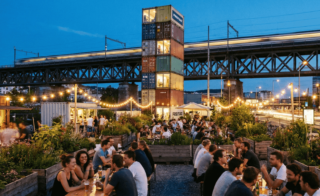 Evening view of Frau Gerolds Garten in Zürich-West featuring the stacked container FREITAG tower and locals socializing under fairy lights near the railway viaduct.