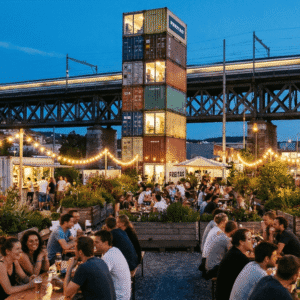 Evening view of Frau Gerolds Garten in Zürich-West featuring the stacked container FREITAG tower and locals socializing under fairy lights near the railway viaduct.