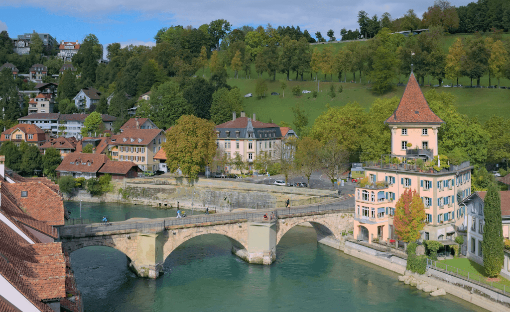 The historic stone arch Untertorbrücke crossing the turquoise Aare River in Bern, Switzerland, featuring the peach-colored Felsenburg tower on the right and the green slopes of the Rosengarten in the background.