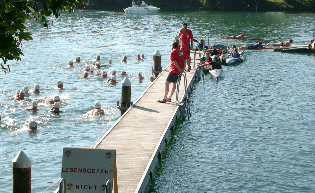 A group of people wearing white swim caps swimming in the green Aare River next to a long wooden pier where staff in red shirts are standing.